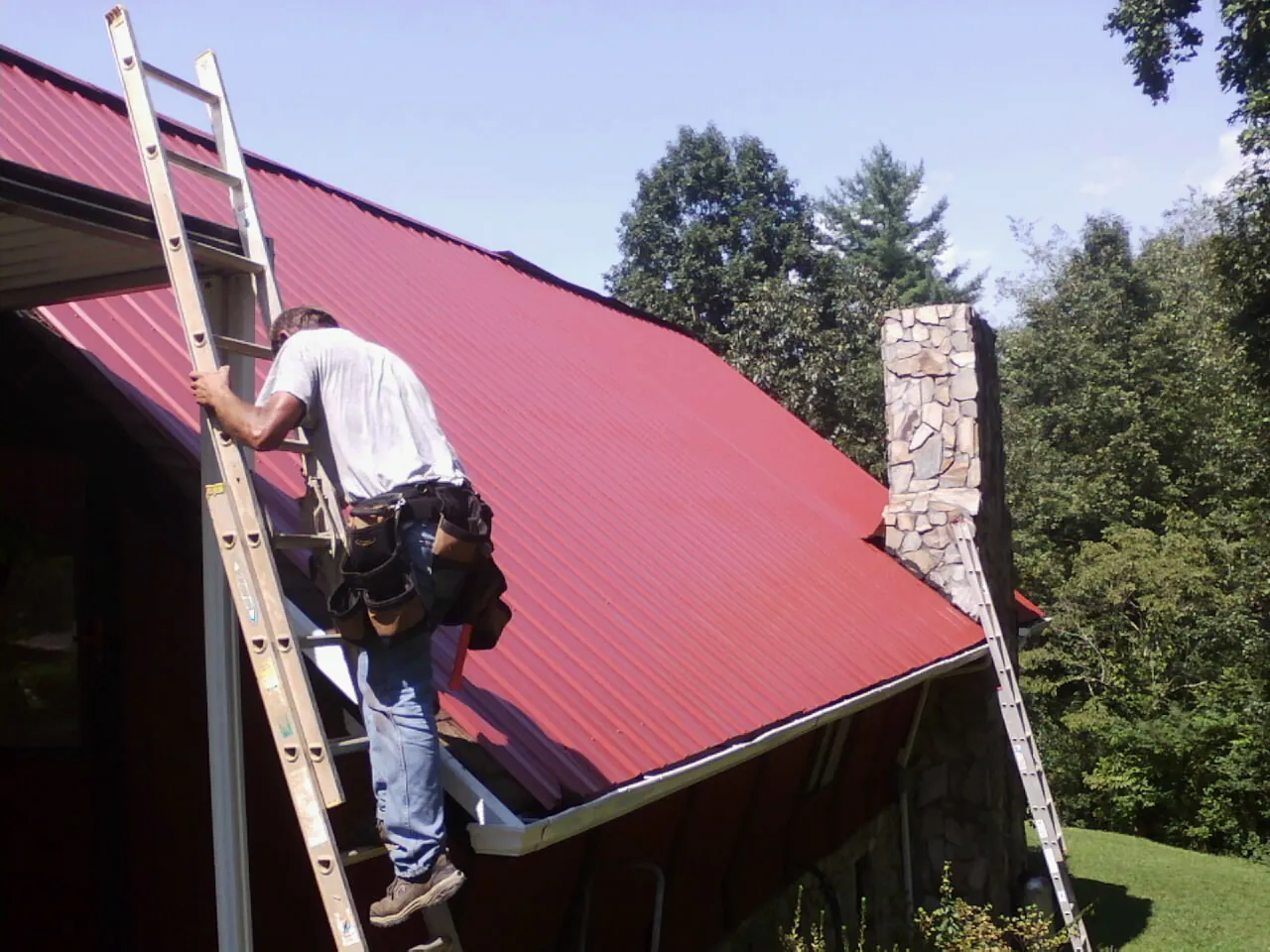 Roofer on a ladder beside a red metal roof and stone chimney — Soffit & Fascia Repair by Roofing Brothers in Montchanin